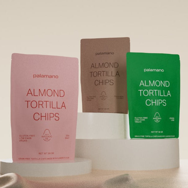 Three Palamano Almond Tortilla Chips pouches. The pouches are in pink (sea salt), brown (cinnamon), and green (lemon), representing different flavor options.